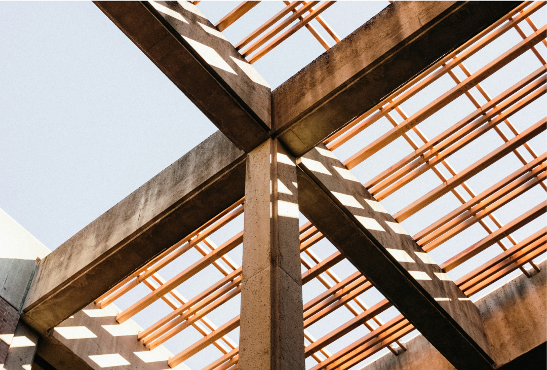 Looking up at intersecting concrete beams and wooden roof framing against a bright sky.