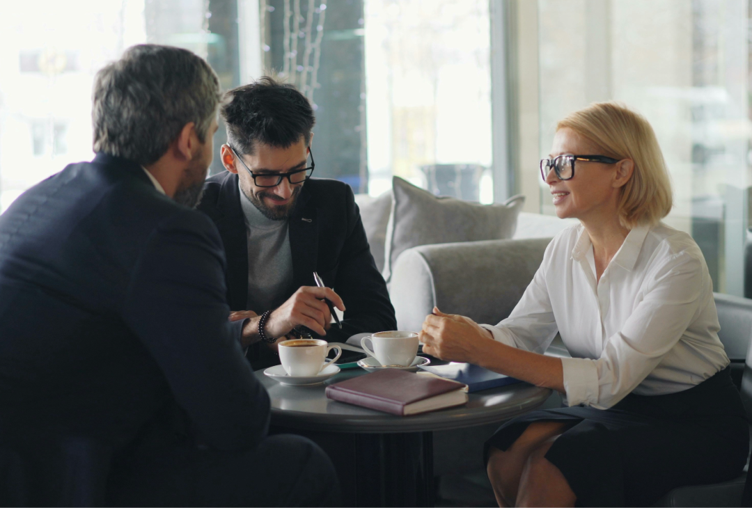 Three business professionals discussing documents over coffee in a modern office, representing collaboration, leadership, and succession planning in a family enterprise.